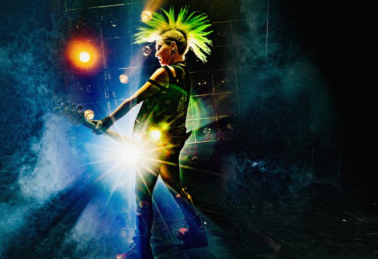 Kuvassa on tummalla näyttämöllä punkkari soittamassa kitaraa ja taustalla on kirkas valo.
