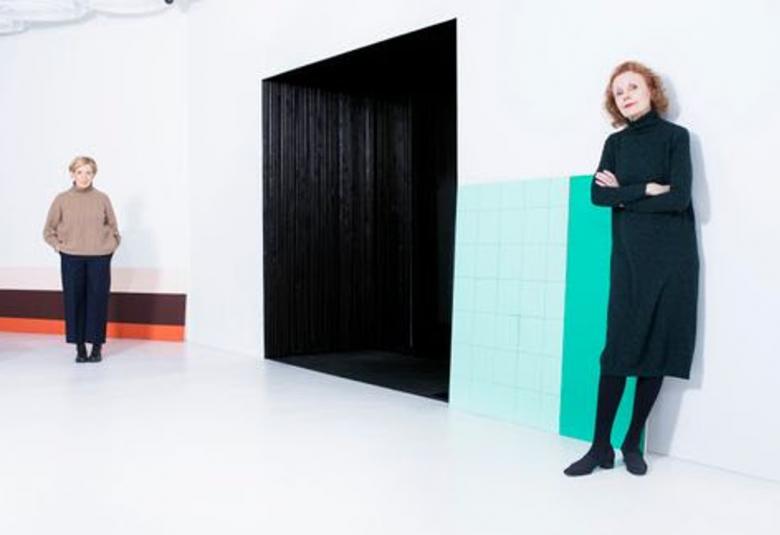 Kuvassa ovat Raija Malka oikealla seinän vierellä takanaan lattialla vaakatasossa olevat palkit päällekkäin.  Ne ovat väriltään oranssi, ruskea ja valkoinen.  Oikealla seisoo Kaija Saariaho vihreän suorakaiteen muotoisen maalipinnan edessä.