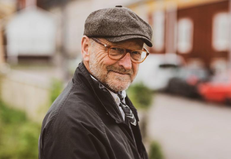 Kuvassa on Tommy Hellsten ulkona Porvoossa lippalakki päässä ja ulkotakissa. Kuvassa on alkusyksy ja takana olevissa pensaissa on vihreät lehdet.