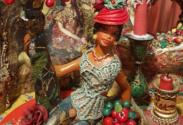 Kuvassa on ruskeapintainen naista esittävä nukke, joka on rehevässä viidakossa ja jossa on puita ja punaisia isoja marjoja.  Nukella on värikäs turbaani päässään.