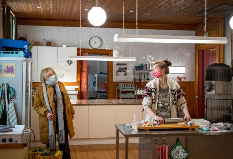 Kuvassa on vasemmalla päällysvaatteissa ja maskissa Leea Klemola ja oikealla leipomassa Emma Kilpimaa.  Kuva on keittiöstä.