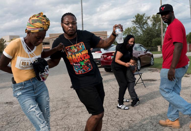 Kuvassa on mustaihoisia henkilöitä. Vasemmalla on hoikka nainen turbaani päässään ja farkuissa vieressään mies mustissa farkuissa ja värikkäässä t-paidassa.  Oikealla on mies, joka on menossa eteenpäin ja katsoo kameraan ja taaempana nainen, jolla on lapsi nojaamassa hänen jalkoihinsa.