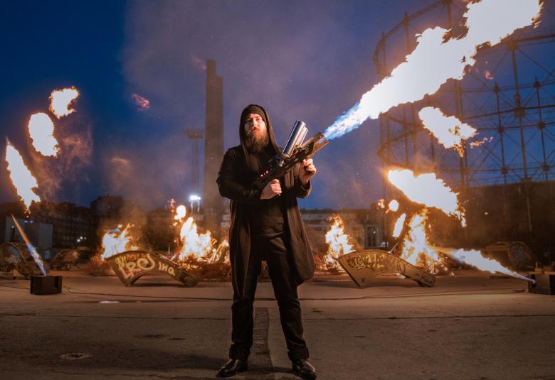 Kuvassa on Rölli Ridanpää iso sytytin pitkällä liekillä etualalla ja taustalla on palava taideteos.  