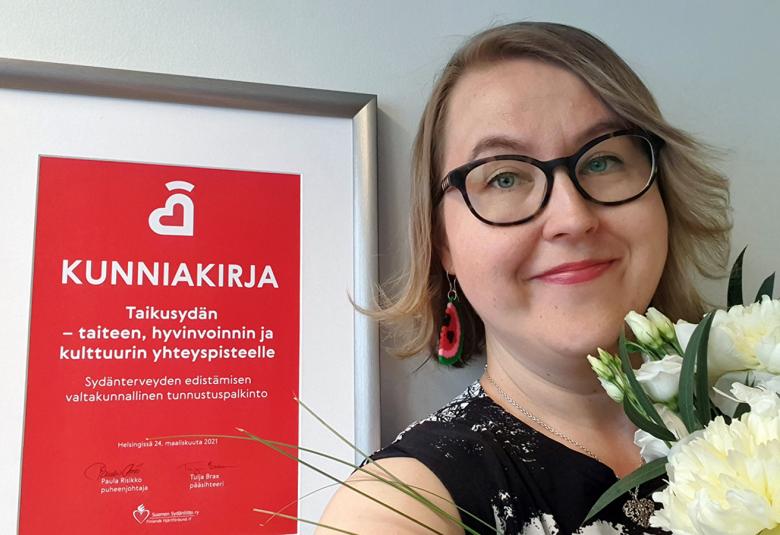 Kuvassa on vasemmalla Taikusydämen kunniakirja ja oikealla on Anna-Mari Rosenlöfin kasvokuva kukat kädessään.