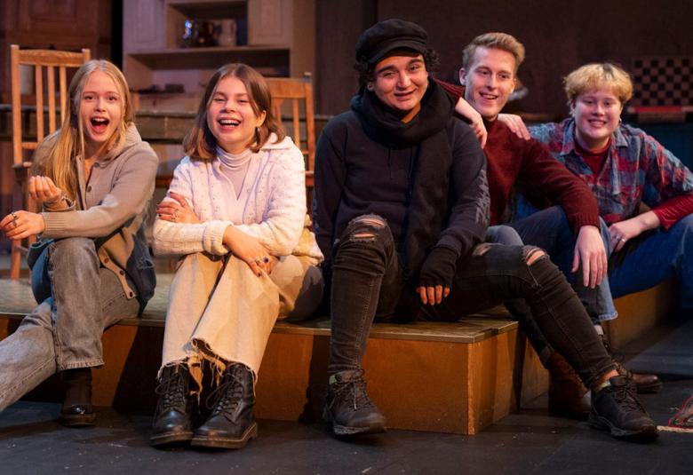 Kuvassa ovat näytelmän nuoret näyttelijät istumassa huonetilassa korokkeella.