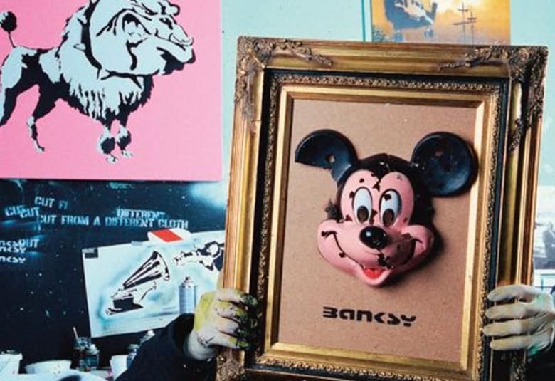 uvassa on Banksyn studio, jossa hän pitää päänsä edessä kullan värisillä raameilla olevaa taulua, jossa on Mikki-hiiren pään kuva.