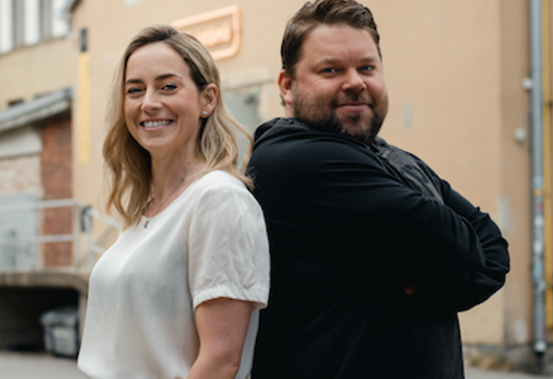 Kuvassa ovat Iina Kuustonen valkoisessa lyhythihaisessa puserossa ja Mikko Töyssy mustassa hupparissa.  He seisovat seläkkäin.  Takana on vaalean ruskea hieman klassisen näköinen entinen teollisuusrakennus.