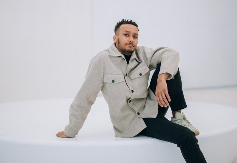 Kuvassa on Ima Iduozee istumassa valkoisen pyöreän lavan päällä nojaamalla vasemmalla kädellään lavaan ja oikea jalka nostettuna myös lavan päälle. Hänellä on vaalean takki päällään.