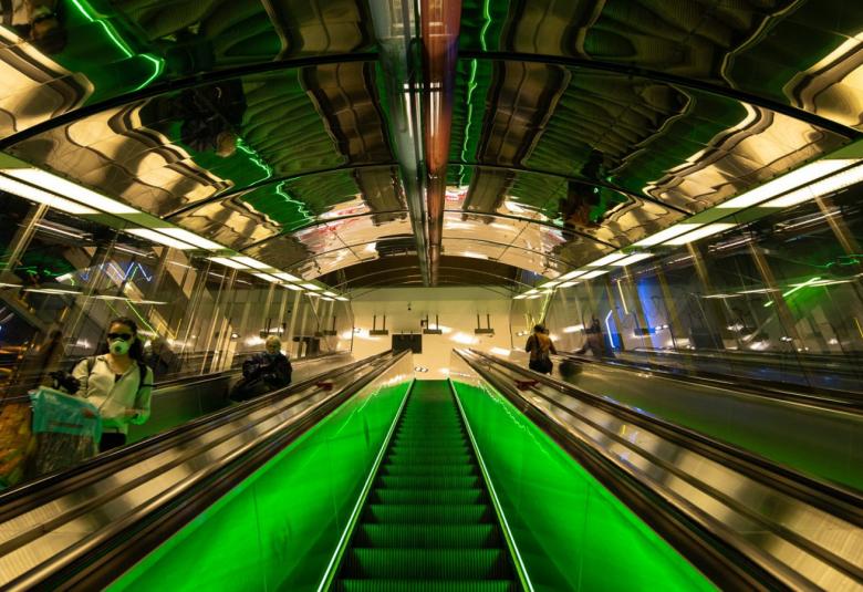 Kuvassa on mediataidekuva metron liukuportaista, jossa on kiiltävät seinät ja katon kuviot.  Keskellä on vihreät leveät raidat ja keskellä yhedet liukuportaat.