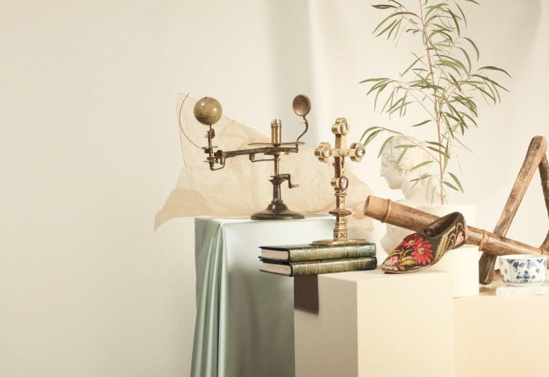 Kuvassa on vasemmalla vaaleaa ja oikealla on esineitä aikaisemmilta historiallisilta ajoilta, mm. koristeellinen suippokärkinen kenkä ja kirjoja.