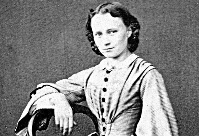 Kuvassa on Minna Canth nuorena istumassa levenevässä mekossa. Kuva on musta-valkoinen.