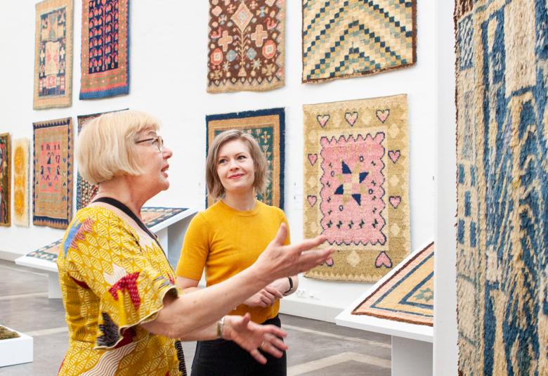 Kuvassa on kaksi keltapuseroista naista esittelemässä valkoisella seinällä olevia erilaisia ja eri kokoisia ryijyjä.