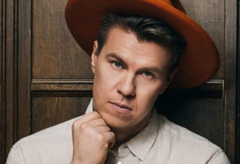 Kuvassa on Olli Halosen kasvokuva.  Hänellä on ruosteen ruskea lierihattu päässään sekä vaalea kauluspaita päällä.