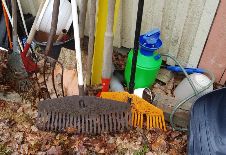 Kuvassa on puutarhatyökaluja kuten haravia, talikko, lapioita, vihreäruiskutuskannu ja muita puutarhatyökaluja nojaamassa vaaleaan seinään. Maassa on ruskeita, kuivia lehtiä.