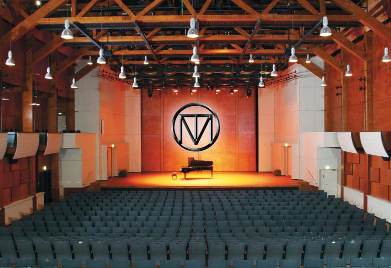 Kuvassa on Savonlinna-sali ja sen katossa puiset orret.  Salissa on tummat tuolit.  Takana on näyttämö, jossa on keskellä flyygeli ja seinällä on m-kirjaimen muotoinen kuvio seinällä.