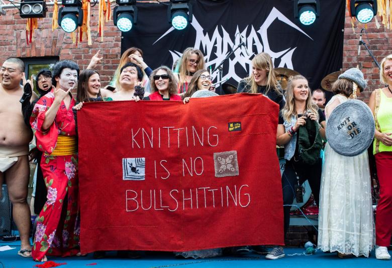 Kuvassa on heavyneulonnan osallistujia, joka ovat punaisen lakanan takana.  Vasemmassa sivussa on muutama japanilainen osallistuja, joista reunassa on sumo-painiasussa oleva mies.  Lakanassa lukee Knitting is no shitting.