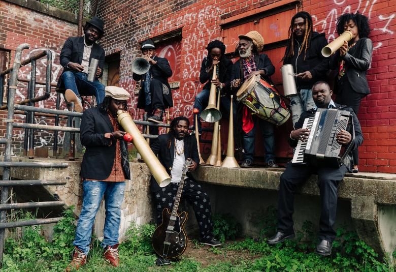 Kuvassa on Lakou Mizikin bändin jäsenet, jotka ovat tiilisen varastorakennuksen edessä lastauslaiturilla ja maassa. Varaston seinässä on graffiteja.  Bändin jäsenillä on esilaisia soittimia kädessään.  Jäsenet ovat kaikki mustaihoisia.