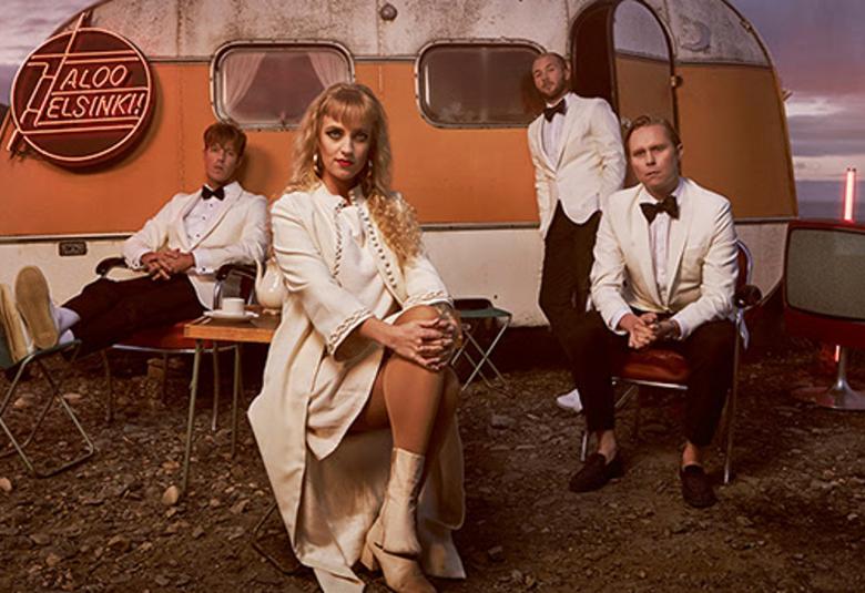 Kuvassa on Haloo Helsinki! istumassa vanhan ruskehtävän asuntovaun edessä ilta-auringon valaistuksessa.