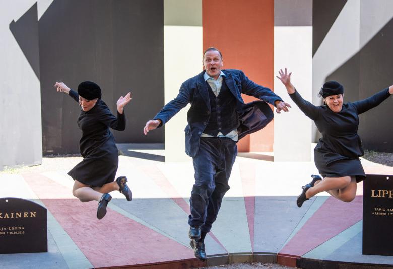 Kuvassa on keskellä miesnäyttelijä ja sivuilla kaksi naisnäyttelijää tanssimassa vauhdikkaasti.  Heillä on kaikilla mustat puvut ja reunoissa on hautakivet.