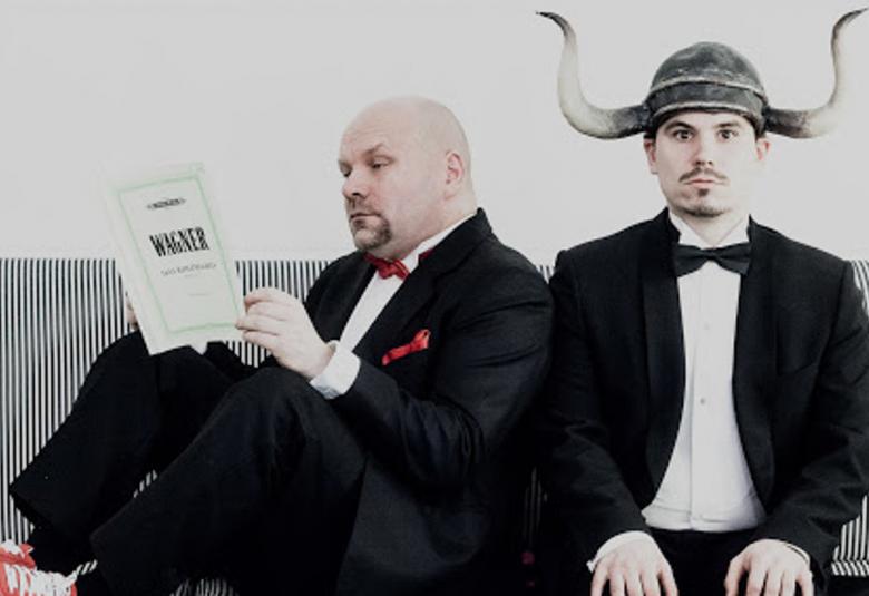 Kuvassa on kaksi The Fabulous Bäckström Brothersin jäsentä istumassa ja toisella on kädessään kypärässä on sarvet. Toinen istuu lukemassa kirjaa vinottain sohvalla jalat myös sohvalla.