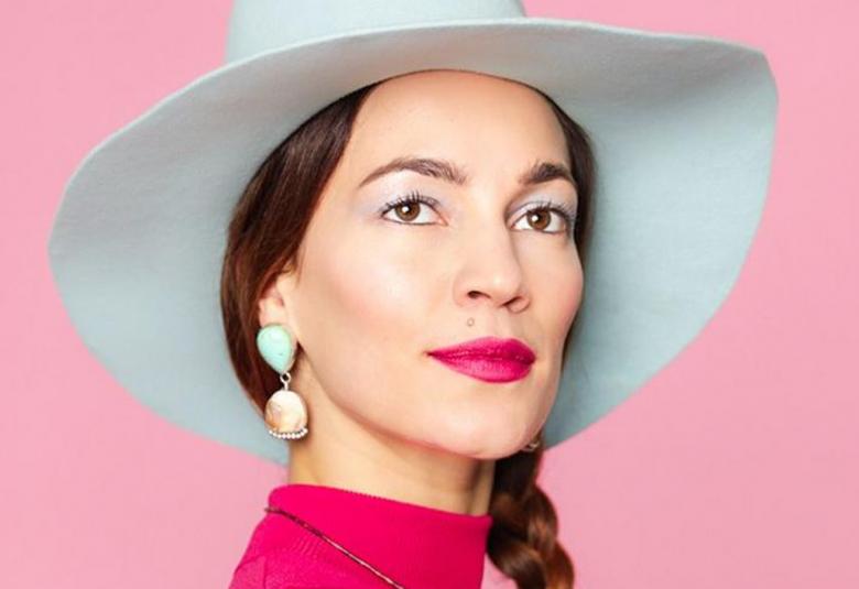 Kuvassa on Meeri Koutaniemi valkoisessa lierihatussa kasvokuvassa.  Tausta on vaalean punainen.