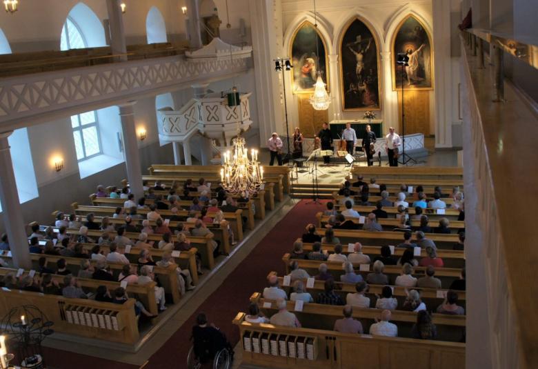 Kuvassa on näkymä Hattulan kirkosta takaparvekkeelta. Kirkossa näkyy penkeissä yleisöä ja taustalla alttaria esiintyjineen.