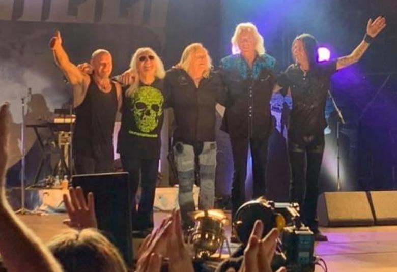 Kuvassa on Uriah Heepin neljä miesesiintyjää esiintymislavalla seisomassa. Taustalla valoja.