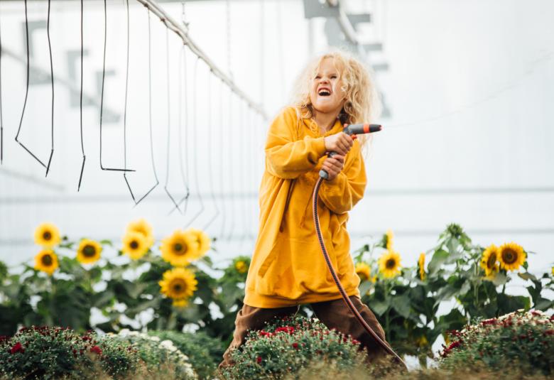 Kuvassa on keskellä Elina Patrakka kasteluletku kädessään seisomassa kasvihuoneessa auringonkukkatarhassa.