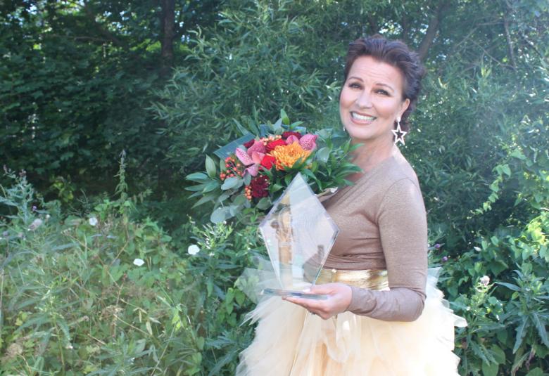 Kuvassa on Arja Koriseva vihreiden pensaiden seassa ja hänellä on vaaleanruskea pusero ja vaalea röyhelöhame.