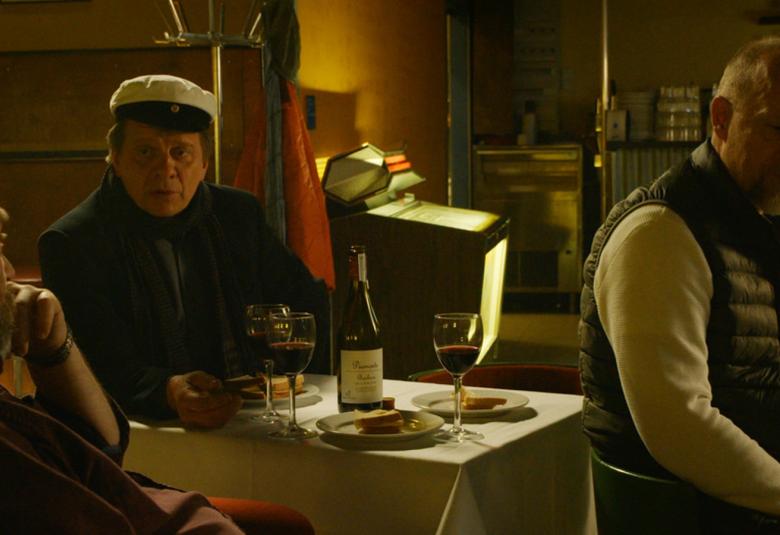 Kuvassa on hämärä baari ja sen pöydässä istuu kaksi ylioppilaslakissa olevaa miestä ja yksi oikealla oleva vinosti tuolillaan.