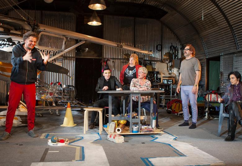 Kuvassa on taustalla pieni lentokone ja vasemmalla Eero Ritala selittämässä innokkaasti.  Oikealla ovat muut näyttelijät istumassa ja katsovat häntä.