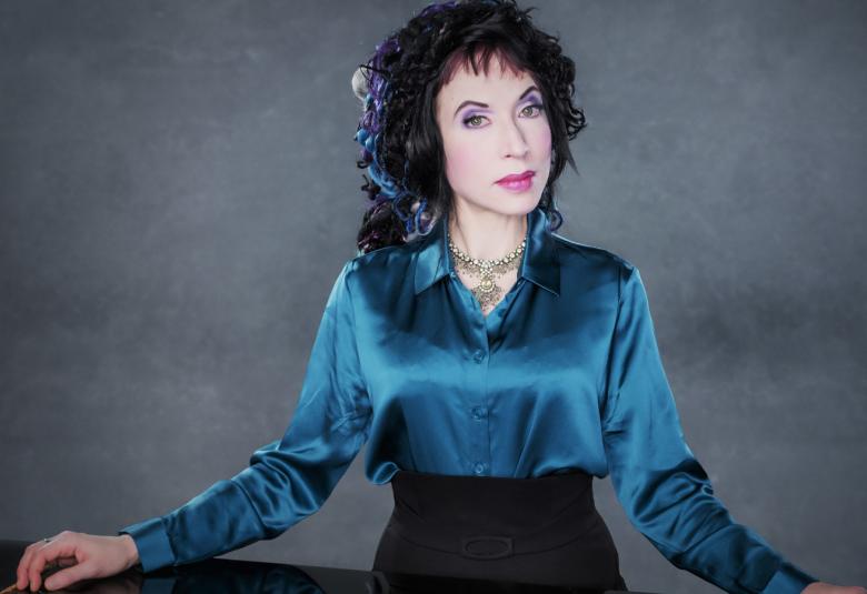 Kuvassa on Sofi Oksanen tummansinisessä kiiltävässä puserossa.  Hänellä on kädet leveästi pöydän pinnalla.