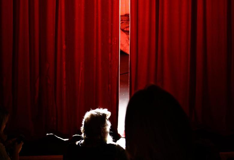 Kuvassa on on punainen esirippu, joka on raollaan ja katsomossa on yksi katsoja.