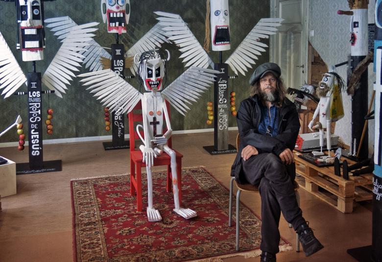 Kuvassa on Jori Tapio Kalliola istumassa tuolilla etuoikealla.  Taustalla on hänen näyttelytilaansa.  Siellä on värikkäitä töitä ja metallisia linnunsiipisiä olentoja keskellä.