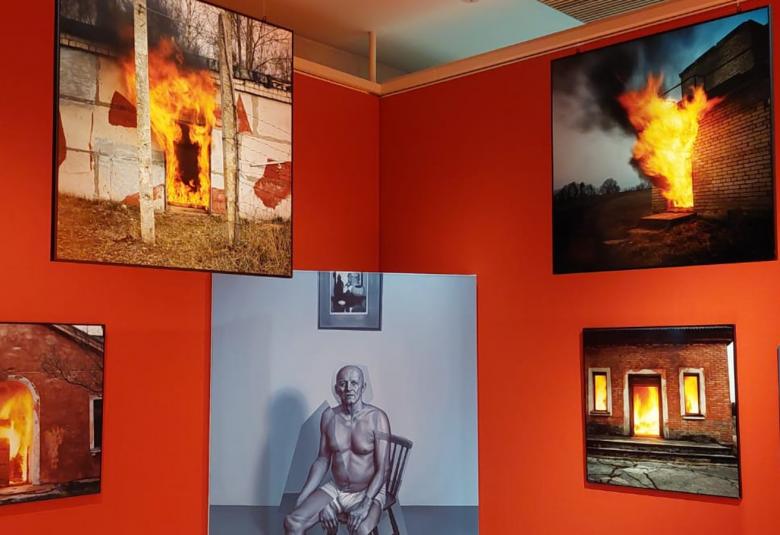 Kuvassa on punaisella seinälle nurkakkain olevilla seinillä erityisesti tulipaloja kuvaavia teoksia ja keskellä on vanhempi mies ilman paitaa.