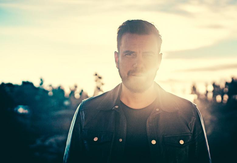 Kuvassa on Antti Ketonen puolivartalokuvassa. Taustalla paistaa aurinko ja luo loistetta Ketosen kasvoille ja värejä taivaalle.