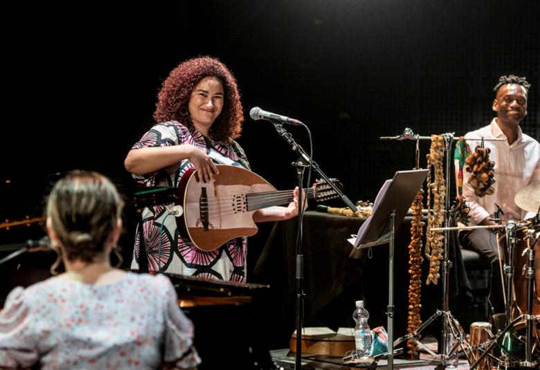 Kuvassa on esiintymislavalla soittamassa tummaa taustaa vasten kitaraan kiharatukkainen nainen ja hänen vieressään mies haitari jaloissaan.  Vasemmalla etualalla selin nainen soittamassa pianoa.