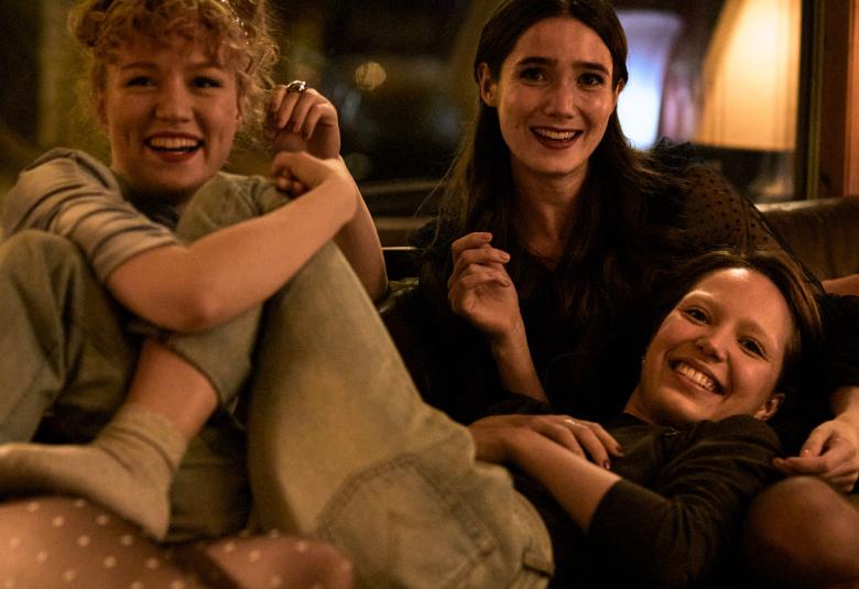 Kuvassa on kolme tyttöä sohvalla ja yksi heistä on makuuasennossa toisten sylissä ja ilmeet ovat iloisia.  Kuvan sävy on ruskea.