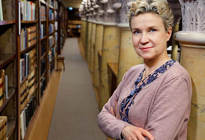 Kuvassa on Sirpa Kähkönen puolivartalokuvassa kuvan oikeassa laidassa kansalliskirjastossa.  Vasemmalla on kirjahyllyrivi ja oikealla pylväitä, joissa on hopeiset kruunukuviot.