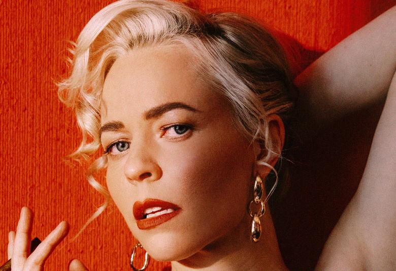 Kuvassa on Paula Vesaan kasvokuva, jossa hän on vaaleahiuksinen. Tausta on punainen.