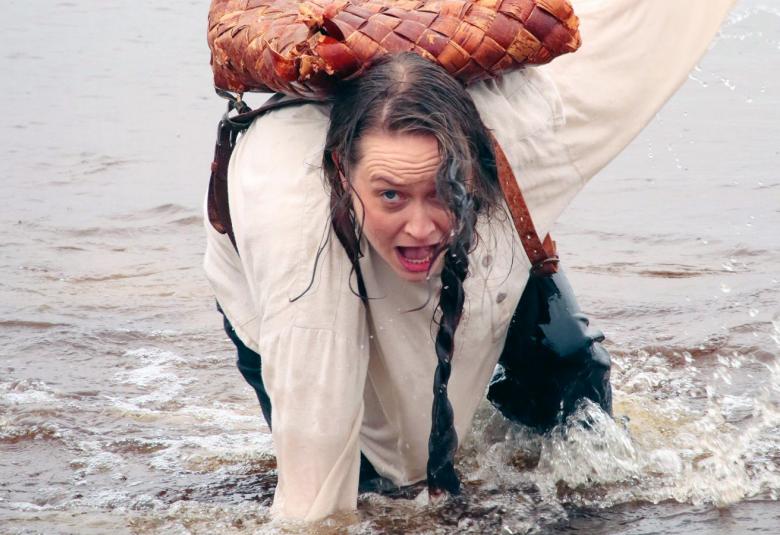 Kuvassa on Vera Veiskola rantavedesä kumartuneena eteenpäin.  Hänellä valkoinen pusero ja hame päällään ja tuohikontti selässään.
