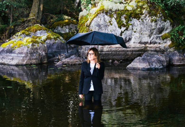Kuvassa on Eija-Liisa Ahtila, joka seisoo tummassa vedessä polvia myöten tummassa pleiserissä ja pitkissä housuissa.  Hän pitää sateenvarjoa kädessään. Takana on kalliota.