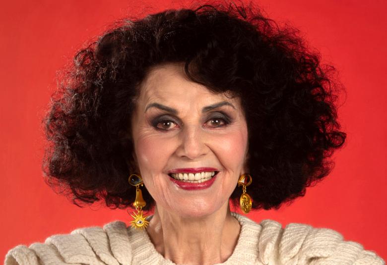 Kuvassa on Lenita Airisto tummassa tukassaan valkoinen villapaita päällään ja takanaan punainen tausta.