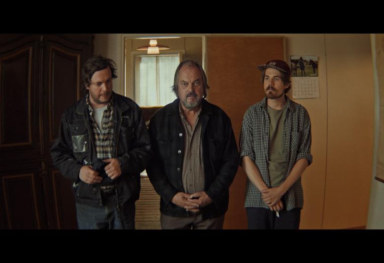 Kuvassa on kolme miesnäyttelijää seisomassa keskellä huonetta.  Kuvan sävy on tumma.