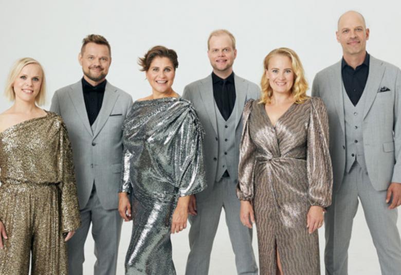 Kuvassa on lauluyhtye Rajaton yhteiskuvassa. Naisilla on kiiltävät puvut ja miehillä harmaat puvut ja mustat paidat.