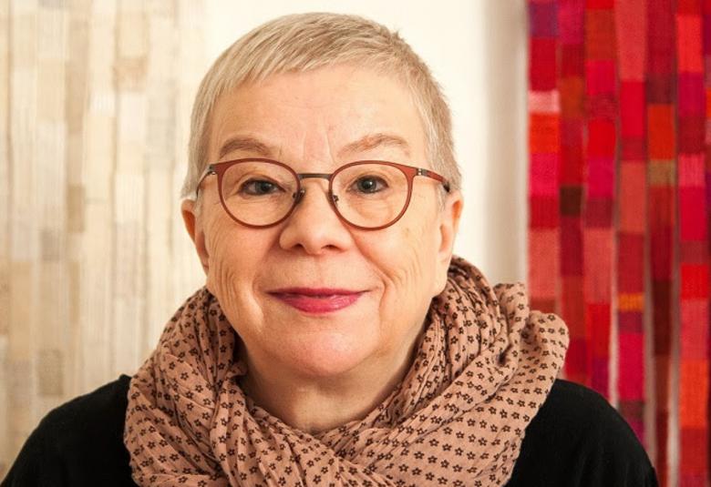 Kuvassa on Riitta Turusen kasvokuva.  Hänellä on vaaleat lyhyet hiukset ja mustasankaiset silmälasit.  Taustalla ovat vasemmalla vaalea ja oikealla punainen tekstiilityöt.