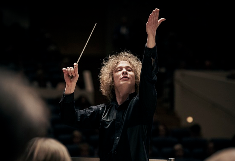 Kuvassa on Santtu-Matias Rouvali johtamassa orkesteria vauhdikkaasti kuvattuna edestä päin tahtipuikko ylhäällä.