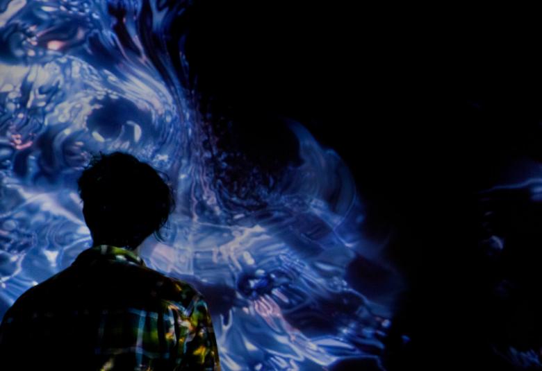 Kuvassa on teos, joka on sininen kiehkuran muotoinen sininen kuvio mustalla pohjalla ja sitä on katselee selin musta hahmo.