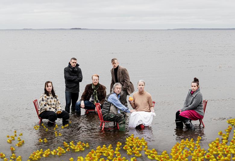 Kuvassa on näytelmän näyttelijöitä istumassa tuoleilla vedesssä puolikaaressa. Edessä ja sivulla kelluu kelstaisia kukkia.  Taustalla näkyy horisontissa mannerta.  Veden väri on harmaa.