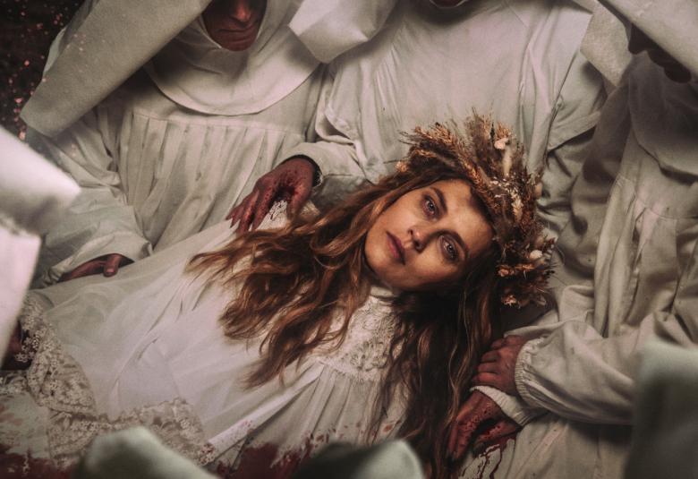 Kuvassa on Teresa Palmer vaaleassa mekossa ja hän on vaaleissa kaavuissa olevien nunnien sylissä.  Nunnien kasvoja ei näy.